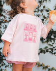 Child wearing a pink t-shirt with a graphic design, standing against a wall with greenery.