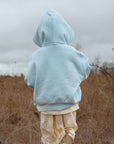 Person wearing a light blue hoodie and beige pants standing in a field with a cloudy sky.
