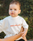 Baby wearing a white shirt with text, held by an adult outdoors.