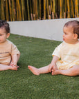 Two babies sitting on grass wearing matching outfits with text.