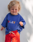 Child wearing a blue shirt with 'PLAY' and red shorts standing against a white wall.