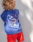 Child wearing a blue sleeveless shirt with a graphic design and red shorts against a white wall.