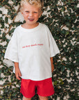 Child wearing a white t-shirt with eat sleep snack repeat and red shorts standing against a leafy background
