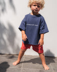 Child wearing eat sleep snack repeat shirt and red shorts standing against a white wall.