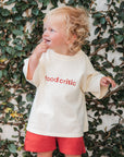 Child wearing a 'food critic' shirt in front of a leafy wall