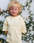 Child wearing a lemon tee with mum needs a margi text, standing against a white wall with green foliage.