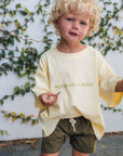 Child wearing a light yellow shirt with text and green shorts standing against a white wall with greenery.