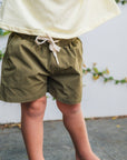 Child wearing green shorts and a yellow top against a white wall with greenery.