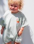 Child wearing a light green organic romper with a peach graphic against a white wall