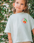 Child wearing a light green family is my world t-shirt with a colorful logo against a leafy background