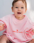 Baby wearing a pink bodysuit with 'Small But Sassy' text sitting on a floor.