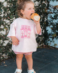 Child eating a donut wearing a pink tee with pink graphics against a leafy background