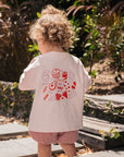 Child wearing a light pink snack club tee with red graphics and shorts, standing on a stone path with plants in the background.