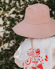 Person wearing a white t-shirt with red graphics and a pink bucket hat, standing against a leafy background.