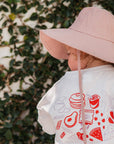 Person wearing a white shirt with red graphics and a pink hat against a leafy background