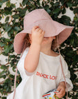 Child wearing a pink sun hat and white dress with 'SNACK LOVER' text, holding snacks against a leafy background.