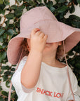 Child wearing a pink sun hat and white dress with 'SNACK LOVER' text, holding snacks against a leafy background.