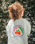 Child wearing a light green t-shirt with a colorful logo and text, standing in front of a leafy green wall.