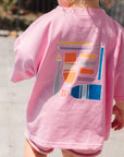 Child wearing a pink shirt with a colorful design on the back, standing outdoors.