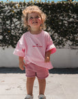 Child wearing small but sassy tee outdoors against a white wall and greenery.