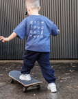 Child wearing a blue t-shirt with white graphics and dark pants, standing on a skateboard.