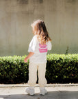 Child wearing a white shirt with a pink design and beige pants standing in front of a concrete wall and green bushes.
