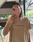 Person wearing a beige t-shirt with 'Mum needs a coffee' text, taking a mirror selfie.