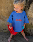 Child wearing a blue shirt with active relaxer red text and red shorts standing against a textured wall.