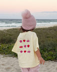 A toddler stands on a sandy beach facing the ocean at sunset, wearing a pale yellow t-shirt with a heart pattern on the back, a pink knit pom-pom beanie, and pink pants.