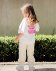 Child wearing a white shirt with text and beige pants standing on a sidewalk.