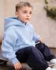Child wearing a light blue hoodie and dark pants sitting on a skateboard.