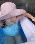 Child in a pool with a large pink hat and blue swim shirt.