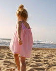 Child in a pink dress standing on a sandy beach looking at the ocean.