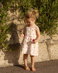 Young girl in a floral dress holding flowers against a stone wall and greenery.