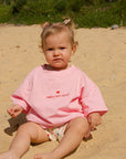Child wearing a pink shirt with text sitting on a sandy beach.