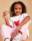 A young girl with curly hair sits on a beige studio background, wearing a white t-shirt with a red heart graphic that reads 'I GOT THIS', light pink pants, white socks, and chunky white sneakers. She is holding a pair of pink-framed sunglasses.