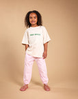 A young girl with curly dark hair stands against a solid beige studio background, wearing an oversized off-white t-shirt with green 'FREE RANGE' text, light pink pants, and pink sandals.