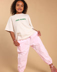 A young girl with curly hair is modeling a cream-colored oversized t-shirt with green 'FREE RANGI' text and light pink baggy pants, standing against a solid beige studio background.