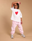 A model posing in a white oversized t-shirt with a red heart graphic 'I got this', light pink pants, and grey sneakers against a solid tan studio background