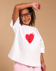 A young girl with curly hair and glasses is modeling a white oversized t-shirt with a red heart graphic that says 'I GOT YOU' in black text, paired with light pink pants, against a solid beige studio background.