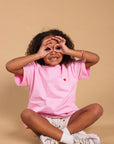 A young child with curly hair sits cross-legged against a solid beige background, wearing a pink t-shirt with a small red heart on the chest, white shorts, and white sneakers.