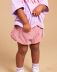 A toddler standing against a beige studio background, wearing an oversized light purple T-shirt with red text, pink and white gingham shorts, and white sneakers.