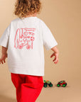 A child seen from behind wearing a white oversized T-shirt with a red graphic print on the back and bright red pants, standing against a plain beige studio background.