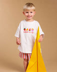 A young blonde boy stands barefoot against a solid beige studio background, wearing a white graphic t-shirt that reads 'SNACK CLUB' with colorful fruit and ice cream illustrations, red and white striped shorts, and holding a large yellow paper airplane.