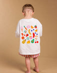 A child standing with their back to the camera, wearing a white t-shirt with a colorful fruit and vegetable pattern on the back, and red, orange, and white striped shorts, against a solid tan studio backdrop.