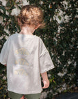 Child wearing a white t-shirt with croissant patterns and green shorts, standing against a leafy background.
