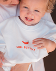 Child wearing a white shirt with red text and a red pepper graphic, held by an adult.