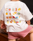 Child wearing a white t-shirt with colorful fruit illustrations and pink shorts.