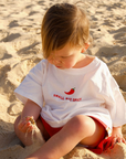 Child sitting on a sandy beach wearing a white t-shirt with text and red shorts.