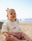 Child sitting on a sandy beach wearing a white shirt with red text and plaid shorts.